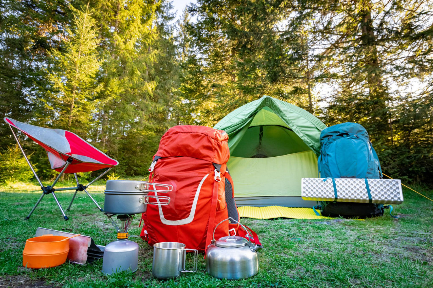 Outdoor camping setup in a forest with a green tent, two large backpacks (one red, one blue), a folding camp chair, metal camping pots, a kettle, and other gear on the grass.